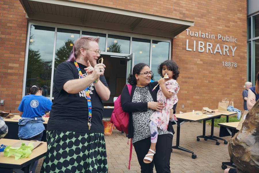 A library staff person, a mother, and a daughter standing and smiling in front of the Tualatin Public Library.