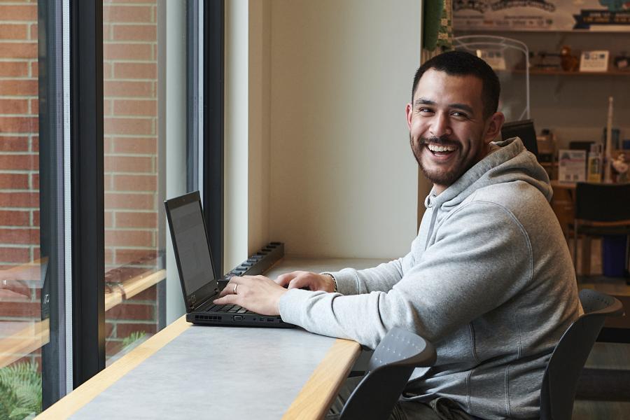 A man using a laptop sits at a table by a window.