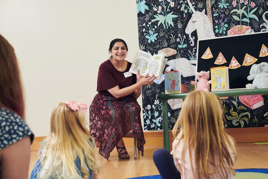 A woman holding a book reads a story to children seated on the floor.