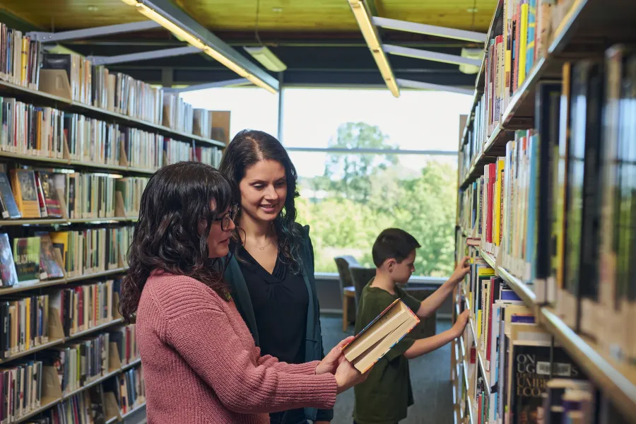 Two women and a young boy looking at books on a shelf in a library.