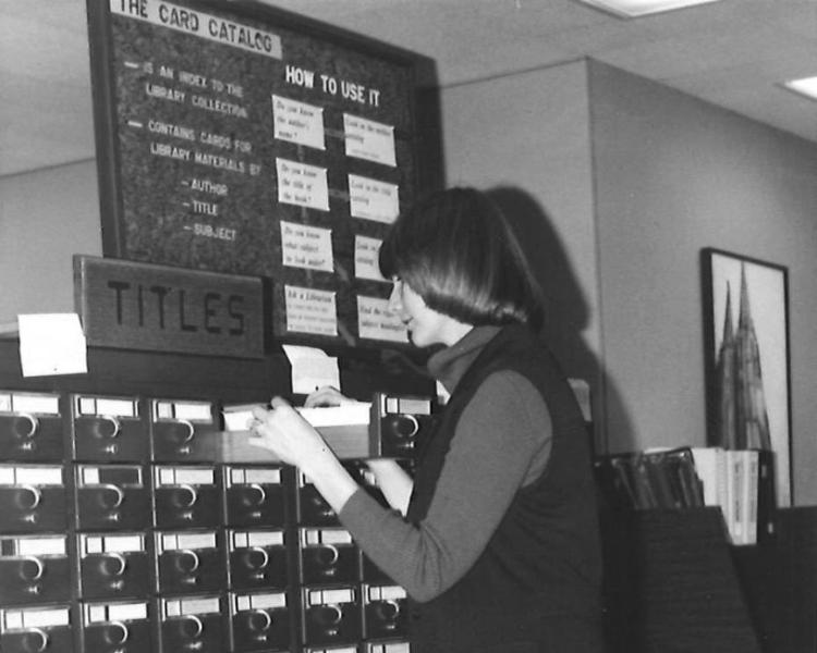 A black and white photo of a woman looking through library cabinet.