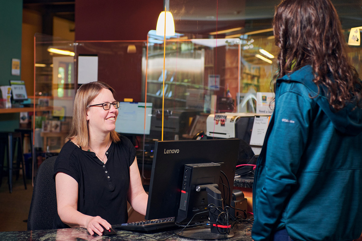 Library staff seated at computer smiling at standing patron.